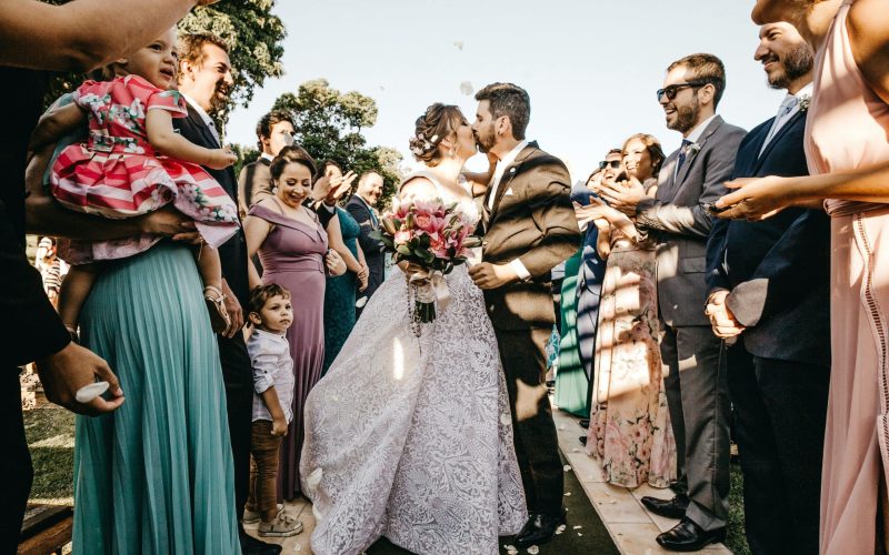 Een bruidspaar kust elkaar aan het einde van een buitenpad terwijl gasten juichen en het begin van hun duurzame relatie vieren. De bruid, die een boeket vasthoudt, draagt een kanten witte jurk en de bruidegom draagt een donker pak. Verschillende aanwezigen omringen hen, waaronder een vrouw die een kind vasthoudt en een jongetje dat omhoog kijkt.