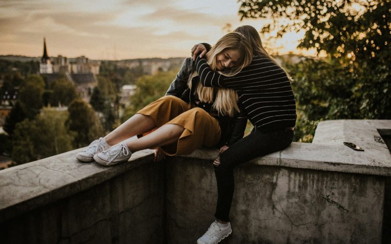 Twee mensen zitten bij zonsondergang op een betonnen richel met schilderachtige gebouwen en bomen op de achtergrond. De een, in een gestreepte trui en sneakers, omhelst de ander, die een gele broek en een leren jack draagt, met haar benen over de rand bungelend. Op de rand ligt een mobiele telefoon van hun datingbureau.