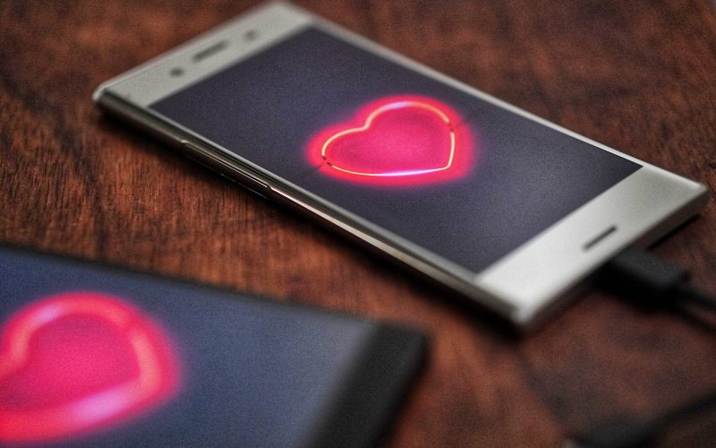 Een smartphone met een gloeiend neonhart op het scherm rust op een houten oppervlak, wat de belofte van een duurzame relatie symboliseert. Een andere smartphone met een wazig scherm is gedeeltelijk zichtbaar op de voorgrond. Beide telefoons zijn verbonden via oplaadkabels.