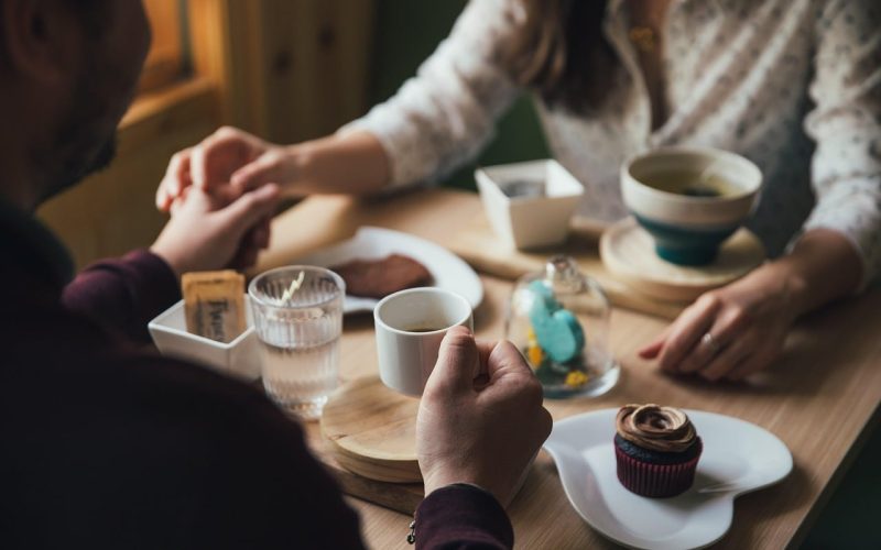 Een stel zit aan een houten tafel, houdt elkaars handen vast en deelt koffie, thee, een cupcake en gebak. De scène voelt warm en intiem aan en biedt inspiratie en datingtips voor wie op zoek is naar gezellige daten-momenten in natuurlijk licht.