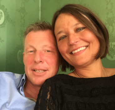 Succesverhalen - Start 6 Een man en een vrouw glimlachen naar de camera terwijl ze een selfie maken. De man, met kort, lichtgekleurd haar, draagt een lichtblauw overhemd. De vrouw heeft bruin haar tot op haar schouders en draagt een zwarte top. Ze lijken samen gelukkig en laten mogelijk hun succes zien via datingbureau-diensten, tegen een achtergrond met een groen patroon.